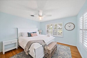 A bedroom with a bed, bedside tables, a ceiling fan, a window with blinds, and a wall clock.