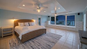 Un dormitorio con una cama grande, un ventilador de techo, una mesita de noche, un ventanal con vistas al mar y un banco empotrado.