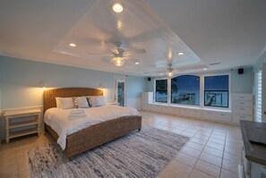 Une chambre dotée d’un grand lit, d’un ventilateur de plafond, d’une table de chevet, d’une fenêtre donnant sur la mer et d’une banquette intégrée.