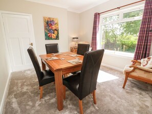 A dining area with a wooden table, black chairs, a painting on the wall, and a window with curtains. - Sundale (Strathaven)