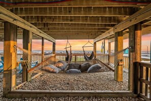 A wooden deck with a hammock and pillows, overlooking a beach with a sunset view. - Beachfront Getaway: Fire Pit, Game Room & Views (Surfside)
