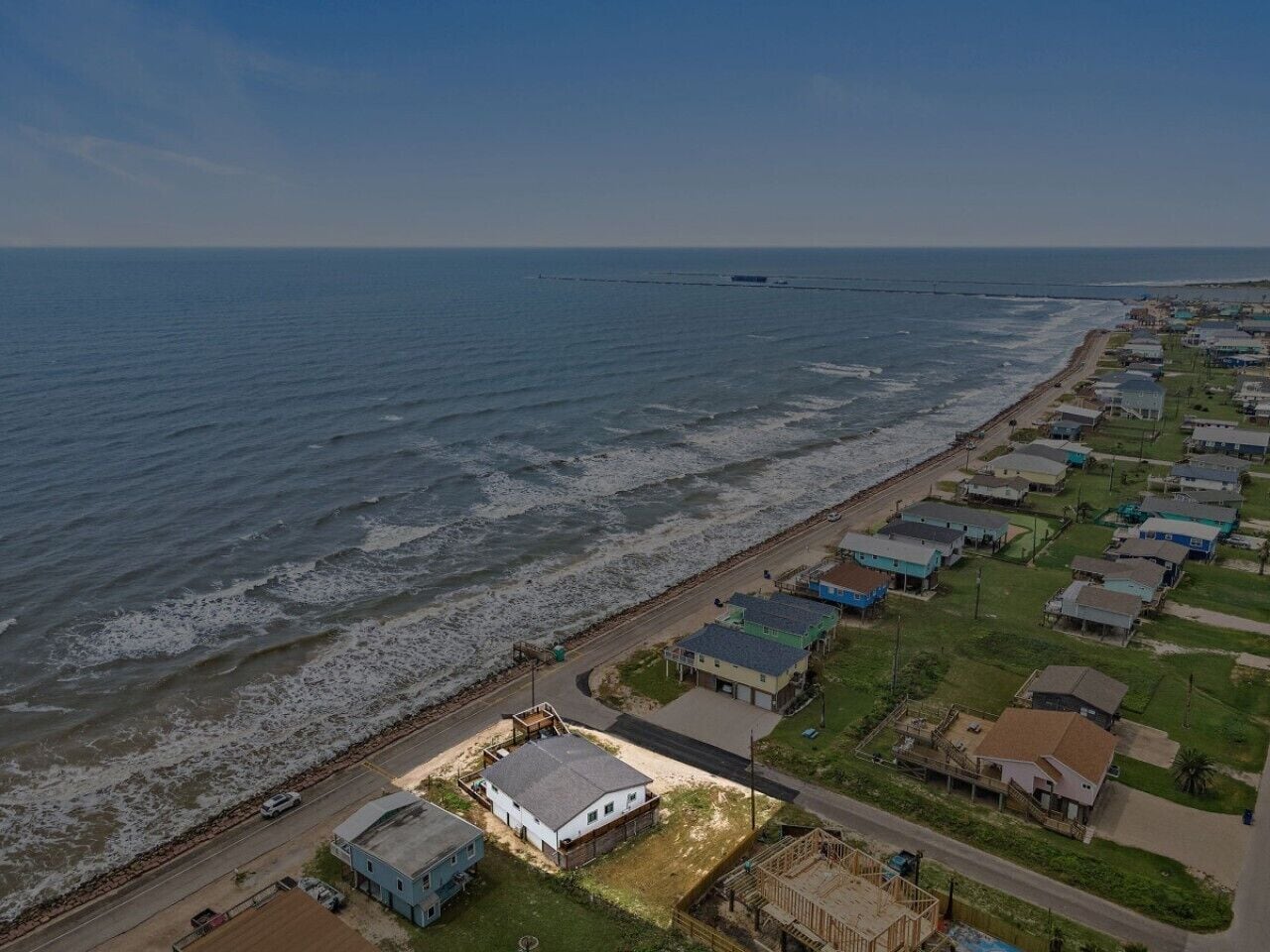 A coastal residential area with houses along the beachfront, a road parallel to the shore, and a distant bridge.