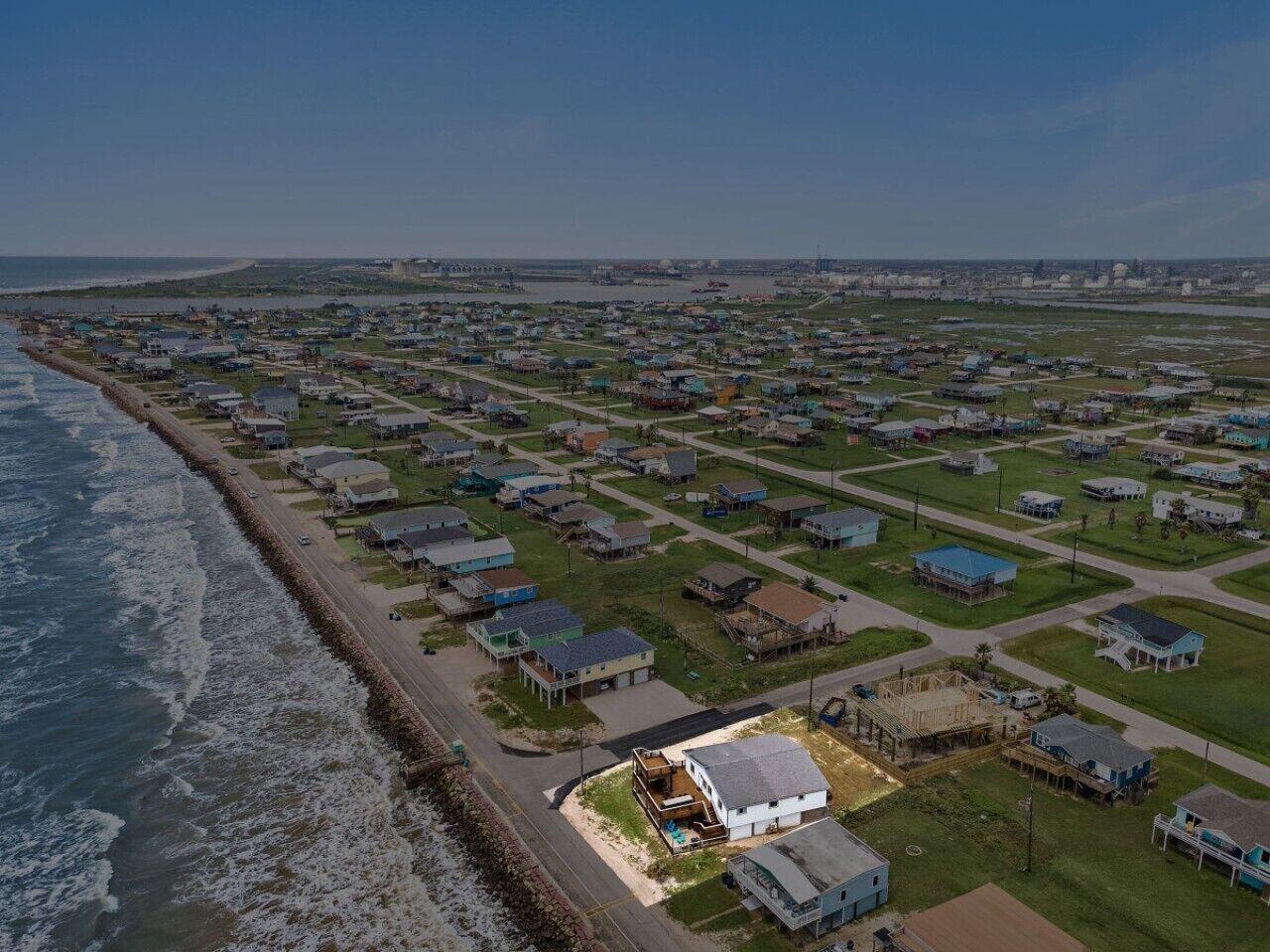An aerial view of a coastal residential area with houses, a beach, and a road parallel to the shore.