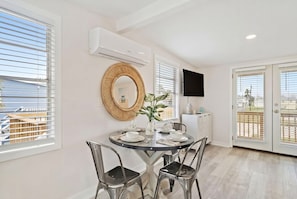 A dining area with a round table, chairs, a wall-mounted air conditioner, a television, and a view through the glass door. - Cozy Beach House w/ Fire Pit, Deck & Game Room (Jamaica Beach)