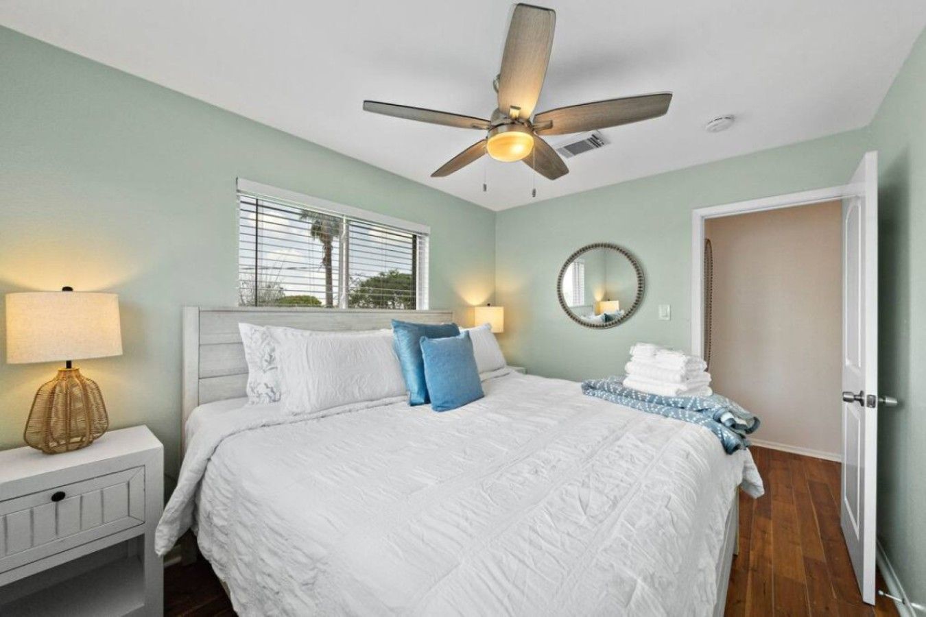 Une chambre à coucher comprenant un lit, un ventilateur de plafond, une lampe de chevet, une fenêtre, un miroir et une porte.