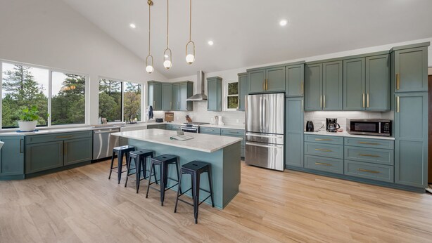 Een moderne keuken met een groot kookeiland, barstoelen, roestvrijstalen apparatuur en kastjes in blauwgroen.