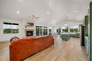 A spacious living room with a fireplace, a large orange sofa, and a view of the outdoors through large windows. - Luxury Home on 5 Acres / Unreal Views! / Hot Tub! (Pagosa Springs)