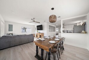 A modern living room with a dining table set for six, a grey sofa, and a kitchen area with a refrigerator.