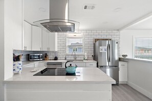 A modern kitchen with white cabinetry, a stainless steel refrigerator, a built-in oven, and a window with blinds. - Oceanview Home & Game Room, Steps to Beach (Galveston)