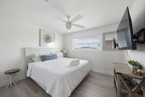 A bedroom with a bed, a ceiling fan, a window with blinds, a bedside table, and a TV mounted on the wall.