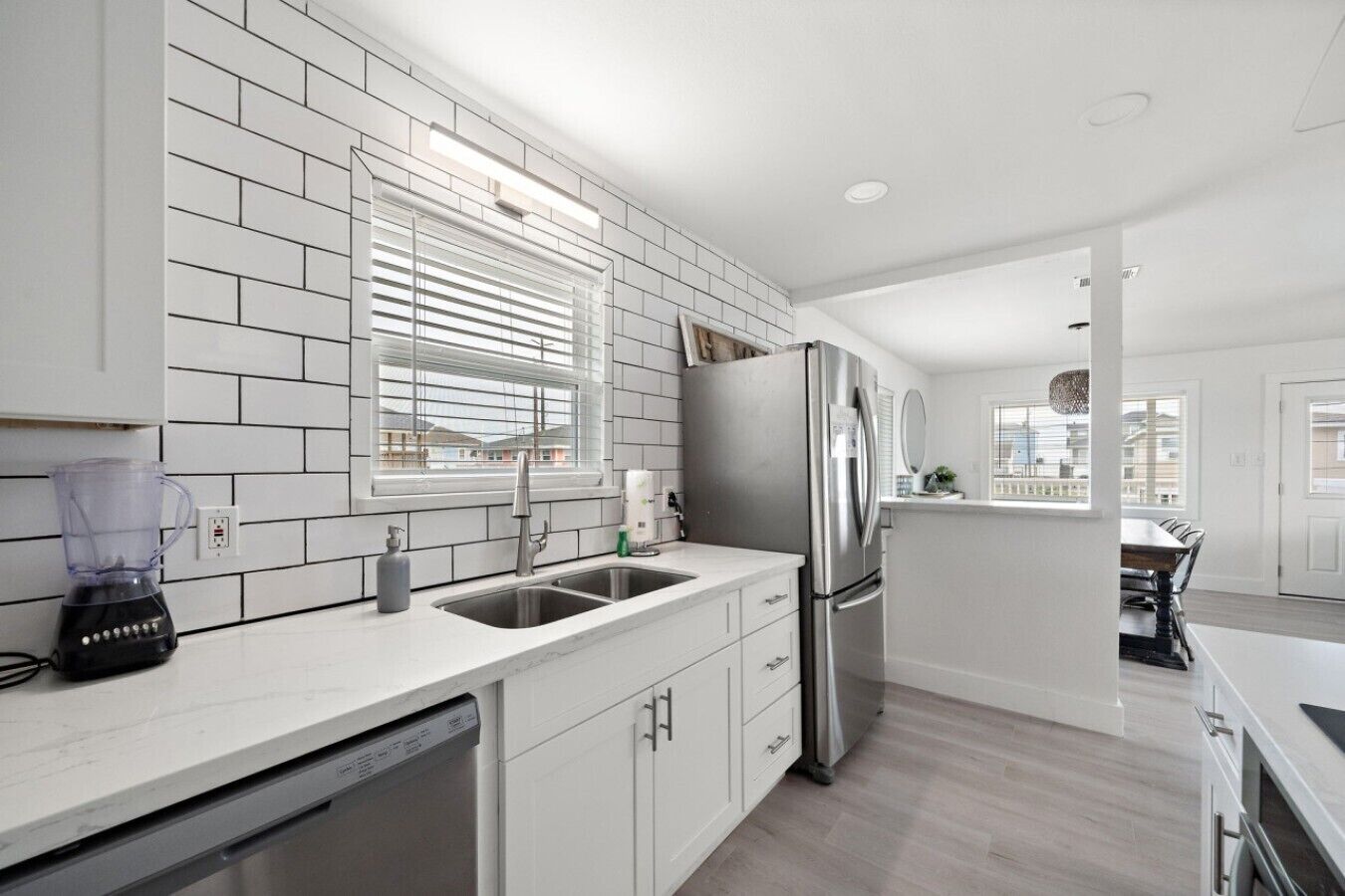 A modern kitchen with white cabinetry, a stainless steel refrigerator, a built-in dishwasher, and a window with blinds.
