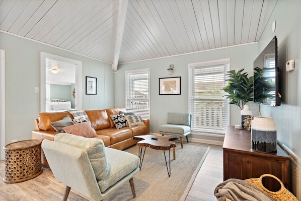 A living room with a brown leather sofa, a light blue armchair, a wooden coffee table, and a TV mounted on the wall. - Charming Canalfront Bungalow with Screened Patio (Galveston)