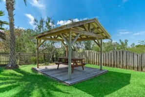 A wooden gazebo with a picnic table, surrounded by a wooden fence and lush green grass. - Texas Vacation Home with Pool, Near the Beach (Galveston)