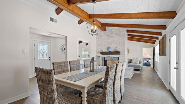 A dining area with a white table and wicker chairs, a brick fireplace, and a hallway leading to a living room.