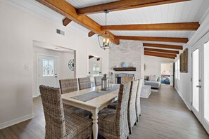 Uma sala de jantar com uma mesa branca e cadeiras de vime, uma lareira de tijolos e um corredor que dá para uma sala de estar.