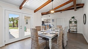 A dining area with a white table set for six, surrounded by wicker chairs, under a wooden beam ceiling with a hanging light fixture.