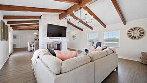 A spacious living room with a large sectional sofa, a fireplace, wooden beams, a flat-screen TV, and a clock on the wall.