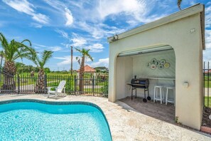 Una piscina, zona de barbacoa cubierta y espacio exterior cercado con palmeras.