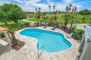 Ein Pool mit geschwungener Kante, umgeben von einer Steindeck, Palmen und einer eingezäunten Liegefläche mit Liegestühlen.