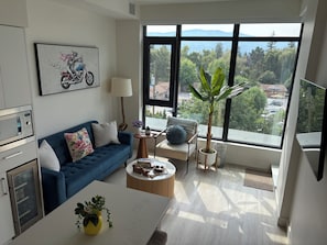 Ein modernes Wohnzimmer mit blauem Sofa, Couchtisch, Fernseher, großem Fenster mit Blick auf Bäume und Häuser sowie einer Küchenzeile mit Mikrowelle und Kühlschrank.