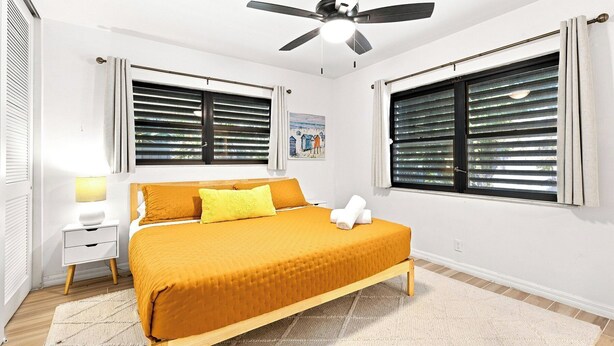 A bedroom with a bed, a ceiling fan, two windows with blinds, a bedside table, and a nightstand. - Heated Pool, Sunny Oasis, 4 bedroom, 9 beds, 4 bathrooms (Fort Lauderdale)