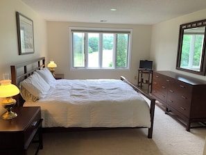 Een slaapkamer met een bed, bedlampen, een dressoir, een televisie en een raam met uitzicht op groen.