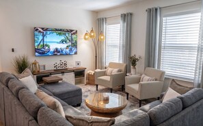 Ein Wohnzimmer mit einer Couch, Sesseln, einem Couchtisch, einem Fernseher und einem großen Fenster mit Jalousien.