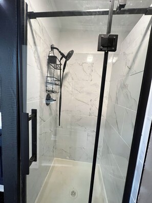 A glass-enclosed shower stall with a marble-tiled wall, a metal rack, and a showerhead.
