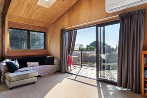A room with a wooden ceiling, a sofa with cushions, a small table, a red chair, and a large sliding glass door leading to an outdoor area. - Roberts Retreat - Auckland Holiday Home (Laingholm)