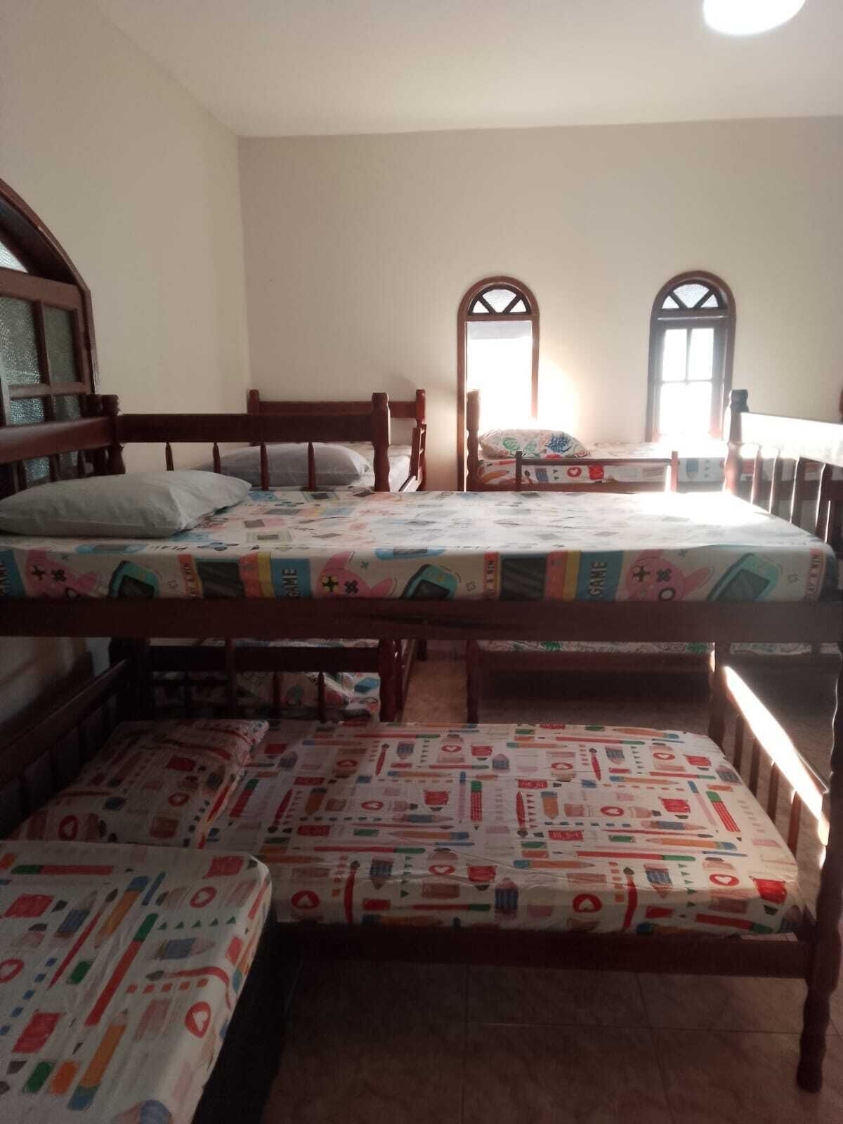 A room with three wooden bunk beds, each with a colorful bedspread, and two arched windows allowing natural light.