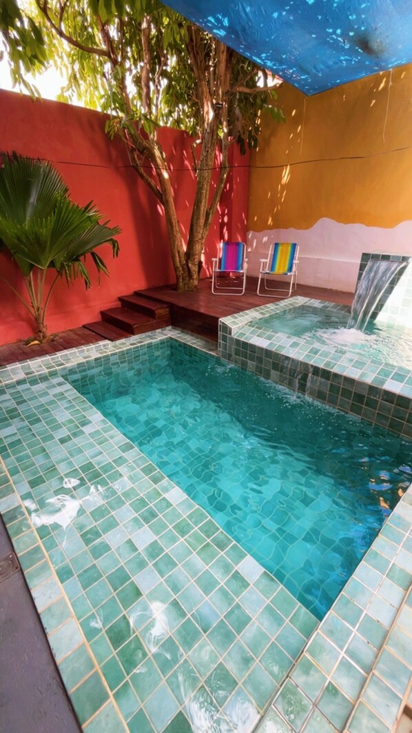 Une piscine dont les bords sont recouverts de carreaux, un arbre et un mur rouge.