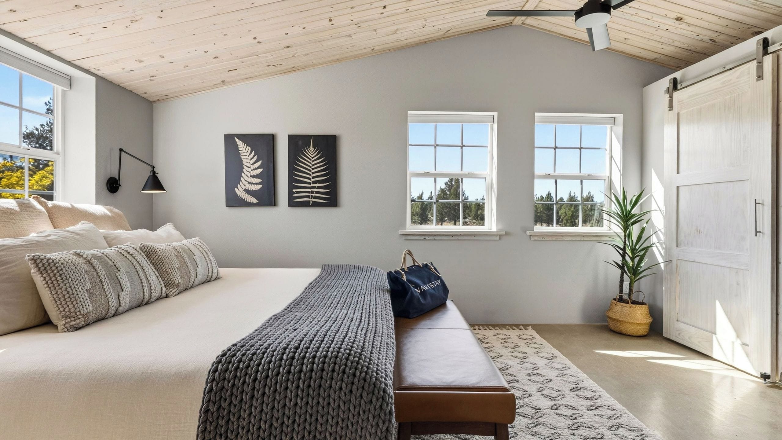 Quarto com cama, um banquinho, duas janelas, ventilador de teto e uma planta.