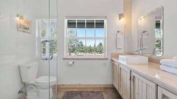 Une salle de bain avec une douche à parois de verre, des toilettes, une fenêtre donnant sur des arbres, un lavabo avec un miroir et un porte-serviettes.