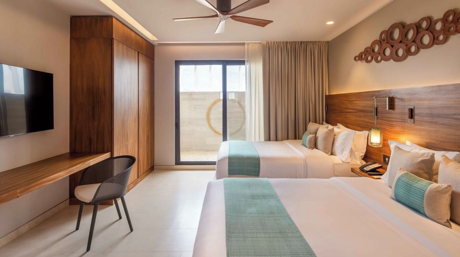 A hotel room with two beds, a desk, a chair, a TV, a ceiling fan, and a large window with curtains.