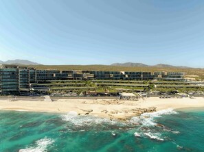 Ein Hotel mit mehreren Stockwerken, ein Strand im Vordergrund und Berge im Hintergrund.