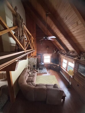 A cozy cabin interior with a stone fireplace, a wooden staircase, a cushioned sofa, and a ceiling fan. - Log Cabin - Hot Tub - Near Kalahari & Camelback (Newfoundland)