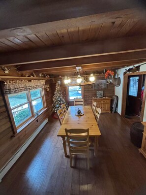 Interior de una cabaña de madera con una mesa de comedor, sillas, un árbol de Navidad y un ventanal grande.