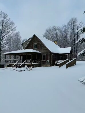 Eine Holzhütte mit Veranda und Stufen, die zu einer Tür führen, umgeben von schneebedeckten Bäumen.