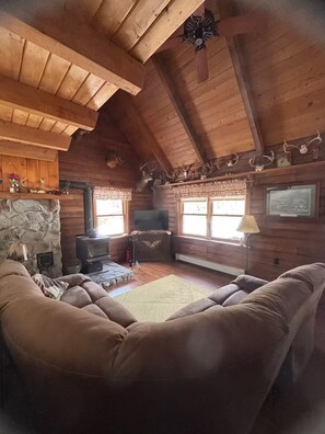 Interior de una cabaña de troncos con chimenea de piedra, vigas de madera en el techo, un televisor y un sofá.