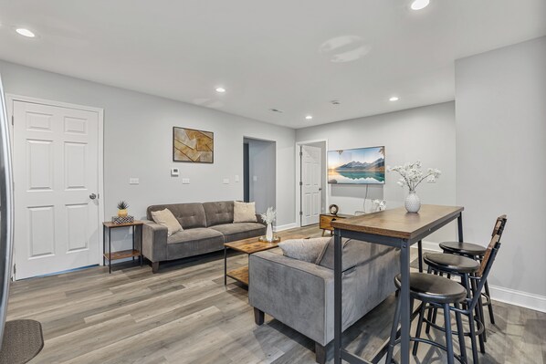 A modern living room with a gray sofa, a coffee table, and a dining area with a wooden table and bar stools.