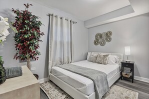 A bedroom with a bed, curtains, a wall art piece, a bedside table with a lamp, and a potted plant.
