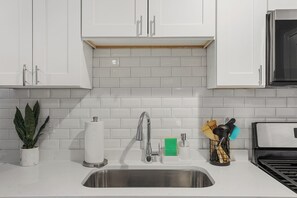 A kitchen with white cabinets, a stainless steel sink, a potted plant, a roll of paper towels, a soap dispenser, and a utensil holder.