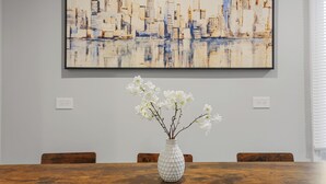 Een eettafel met een witte vaas vol bloemen, een schilderij van een stadsgezicht in een lijst aan de muur, en houten stoelen.