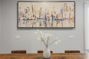 A dining table with a white vase of flowers, a framed cityscape painting on the wall, and wooden chairs.