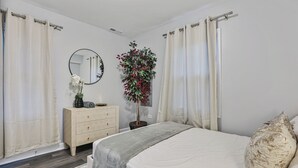 Een slaapkamer met een bed, een kaptafel, een spiegel, een kamerplant en gordijnen.