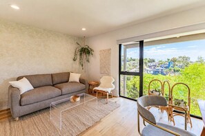 Ein modernes Wohnzimmer mit Sofa, Couchtisch und Sesseln, große Fenster mit Blick auf Häuser und Grünflächen.