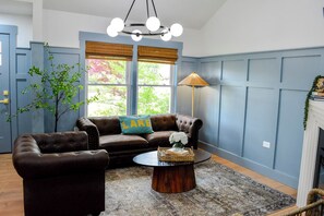Ein Wohnzimmer mit einem braunen Ledersofa, einem Couchtisch mit Korb und Blumen, einer Lampe und Blick auf Bäume durch große Fenster.