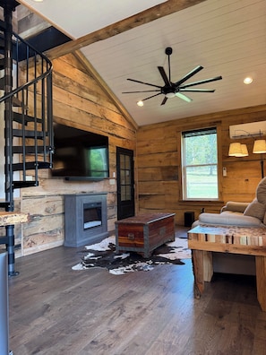 A rustic cabin interior with a fireplace, wooden walls, a ceiling fan, and a cozy seating area.