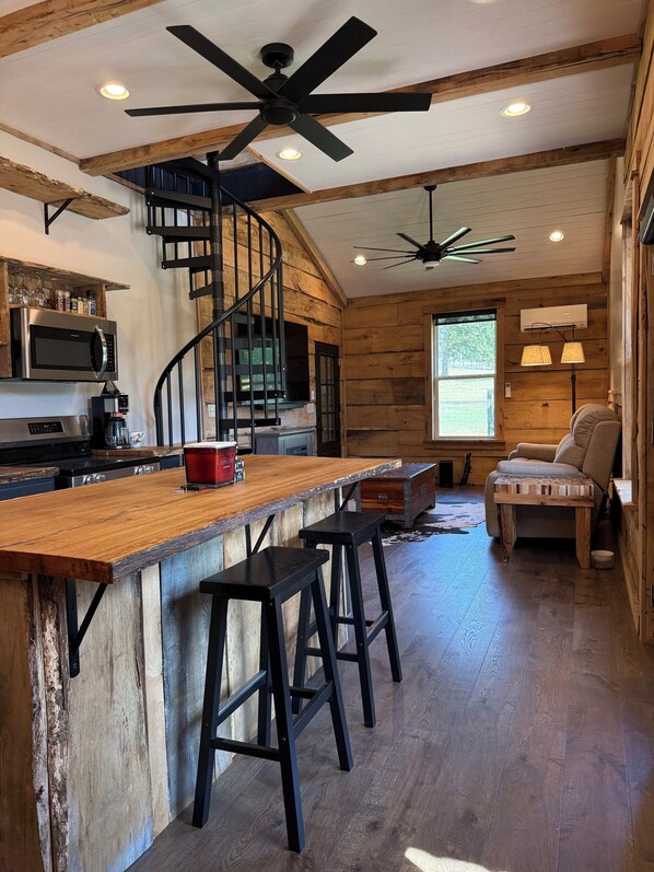A rustic kitchen with a wooden bar, bar stools, a spiral staircase, and a ceiling fan.