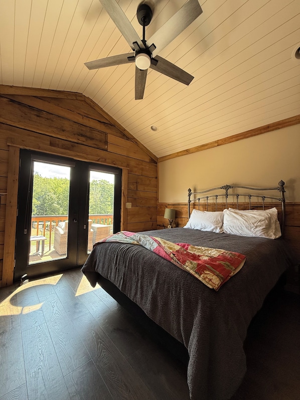 A bedroom with a bed, a ceiling fan, a wooden ceiling, and a view through a sliding door.